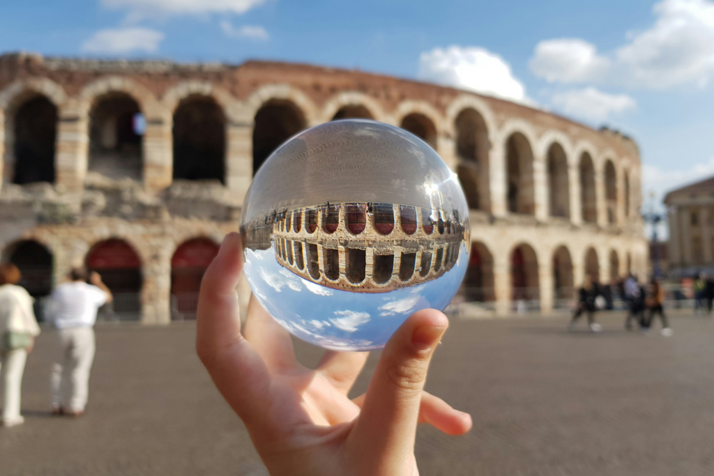 Una mano tiene una pallina in vetro davanti all'Arena di Verona, creando così un riflesso al contrario di essa.